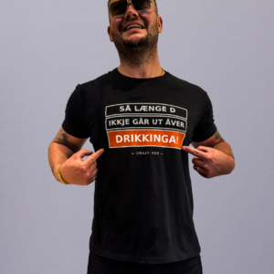 Smiling man in black T-shirt and sunglasses points at the bold orange word 'DRIKKIGA!' on his shirt against a gray background.