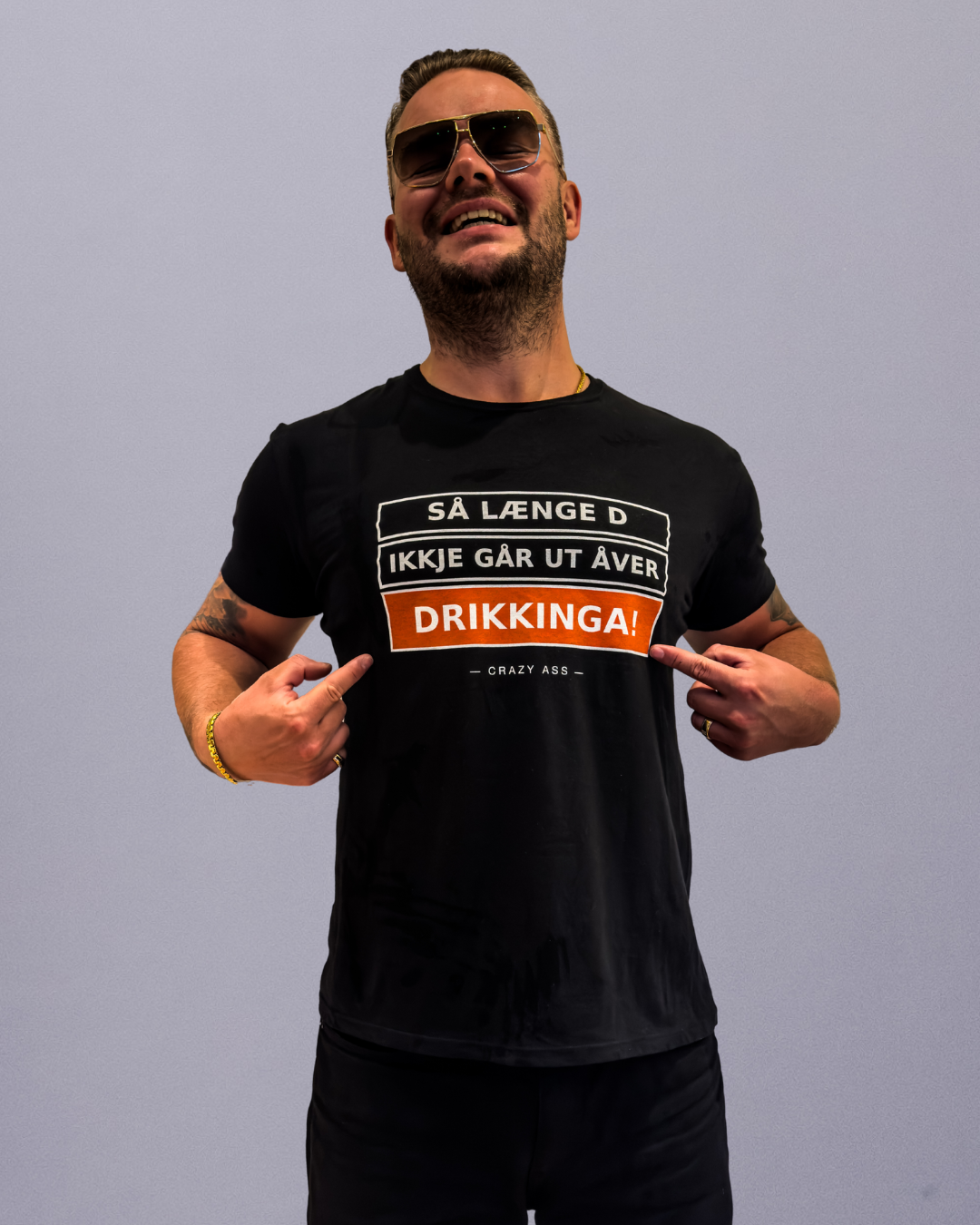 Smiling man in black T-shirt and sunglasses points at the bold orange word 'DRIKKIGA!' on his shirt against a gray background.